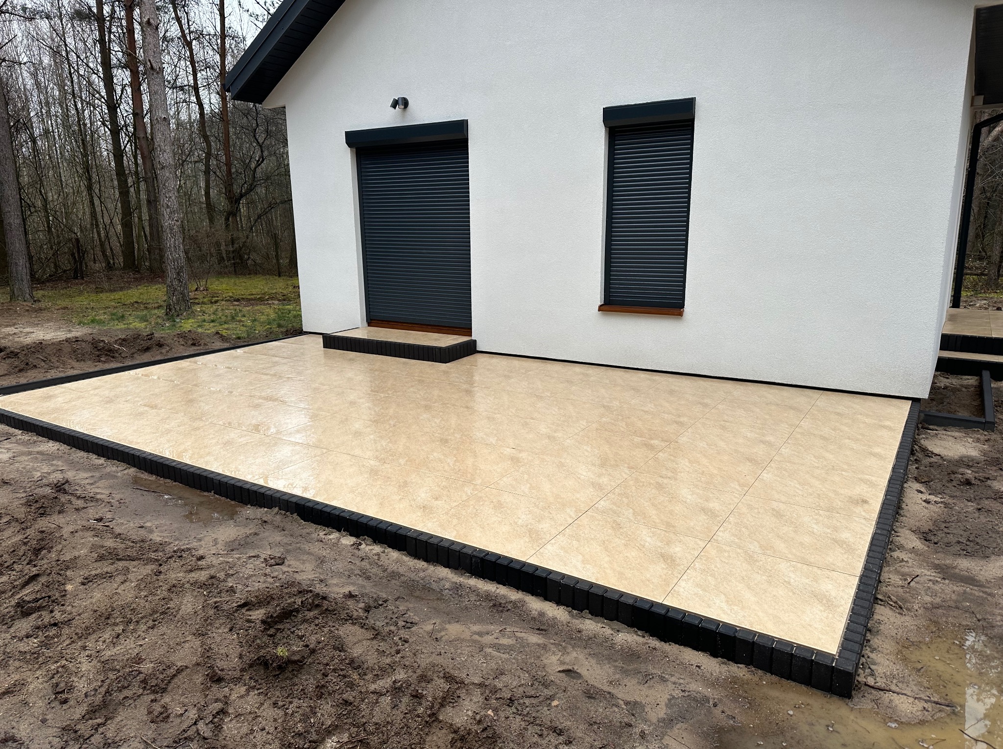 Jasna, kwadratowa kostka na tarasie przy białym domu z szarymi roletami. Czarny obrzeże kostki. Widoczne drzewa w tle. Ukończony projekt.