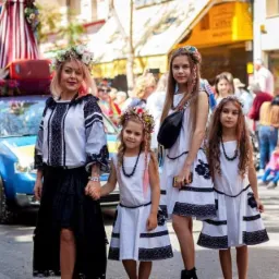 Kobieta z r&oacute;żowymi włosami i trzy dziewczynki w białych sukienkach z czarnymi haftami, wianki na głowach, stoją na ulicy podczas festynu.