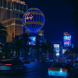 Tobiasz Brakoniecki - Nocny widok na Las Vegas Strip z neonami hoteli Planet Hollywood i Paris, z charakterystyczną wieżą Eiffla w kształcie balonu, ruch uliczny z rozmazanymi światłami samochodów, palmy i jasne reklamy.