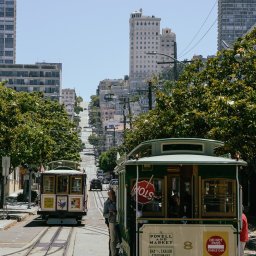 Tobiasz Brakoniecki - Dwa zielone tramwaje linowe w San Francisco, jeden z napisem 'Powell and Market' i numerem 8, jadące po torach w dół ulicy z wysokimi budynkami i stromym wzniesieniem w tle.