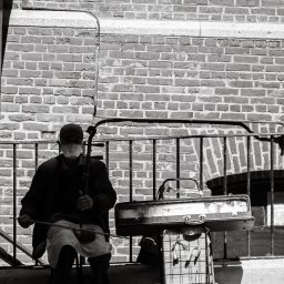 Tobiasz Brakoniecki - Czarno-białe zdjęcie ulicznego grajka z Poznania, siedzącego na krześle z chińskimi skrzypcami erhu, na tle ceglanej ściany i metalowej barierki.