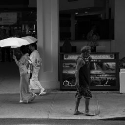 Tobiasz Brakoniecki - Czarno-białe zdjęcie przedstawia dwie kobiety w kimonach z parasolkami idące chodnikiem obok mężczyzny jedzącego coś, na tle punktu informacyjnego z plakatami o atrakcjach turystycznych.
