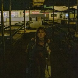 Tobiasz Brakoniecki - Portret kobiety z ciemnymi włosami i mocnym makijażem oczu w opuszczonej hali targowej z metalową konstrukcją dachu i pustymi stoiskami w tle, nocne oświetlenie.