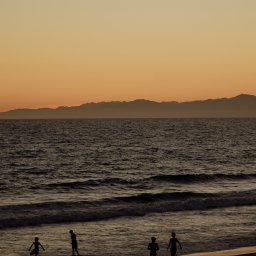 Tobiasz Brakoniecki - Sylwetki czwórki dzieci bawiących się w morzu o zachodzie słońca, w tle zarys górski pod pomarańczowym niebem.