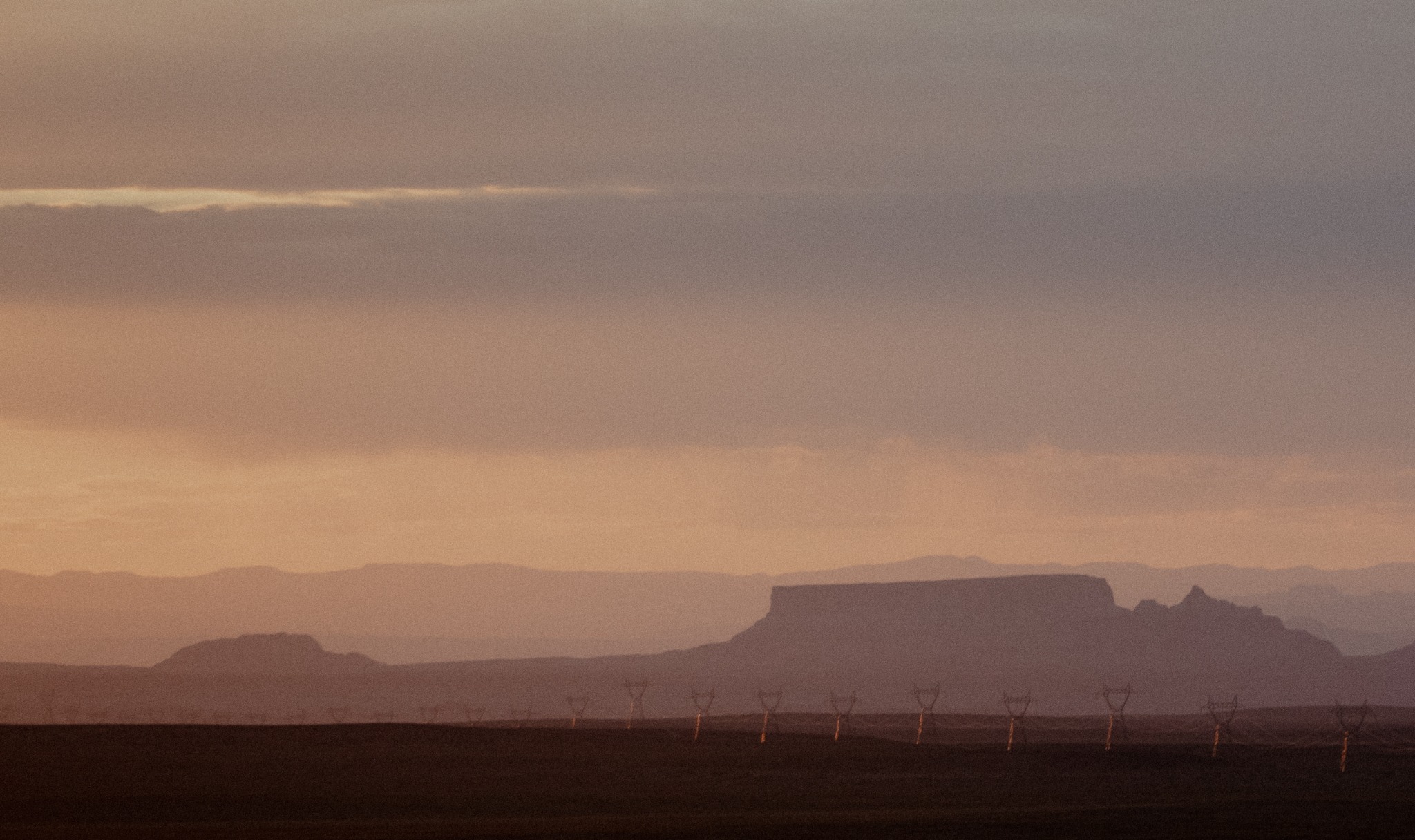 Silhouetted mesas and mountains fade into the distance under a hazy, peach-colored sky, with a row of power line towers sparsely dotting the foreground.