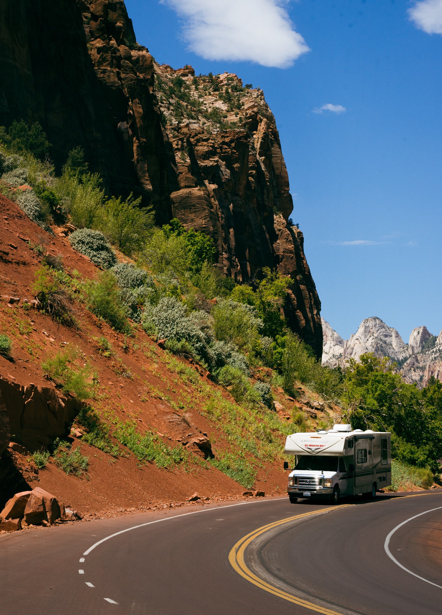 Biały kamper El Monte RV na zakręcie asfaltowej drogi z podwójną żółtą linią, w tle czerwona góra porośnięta zielenią i błękitne niebo z chmurami.