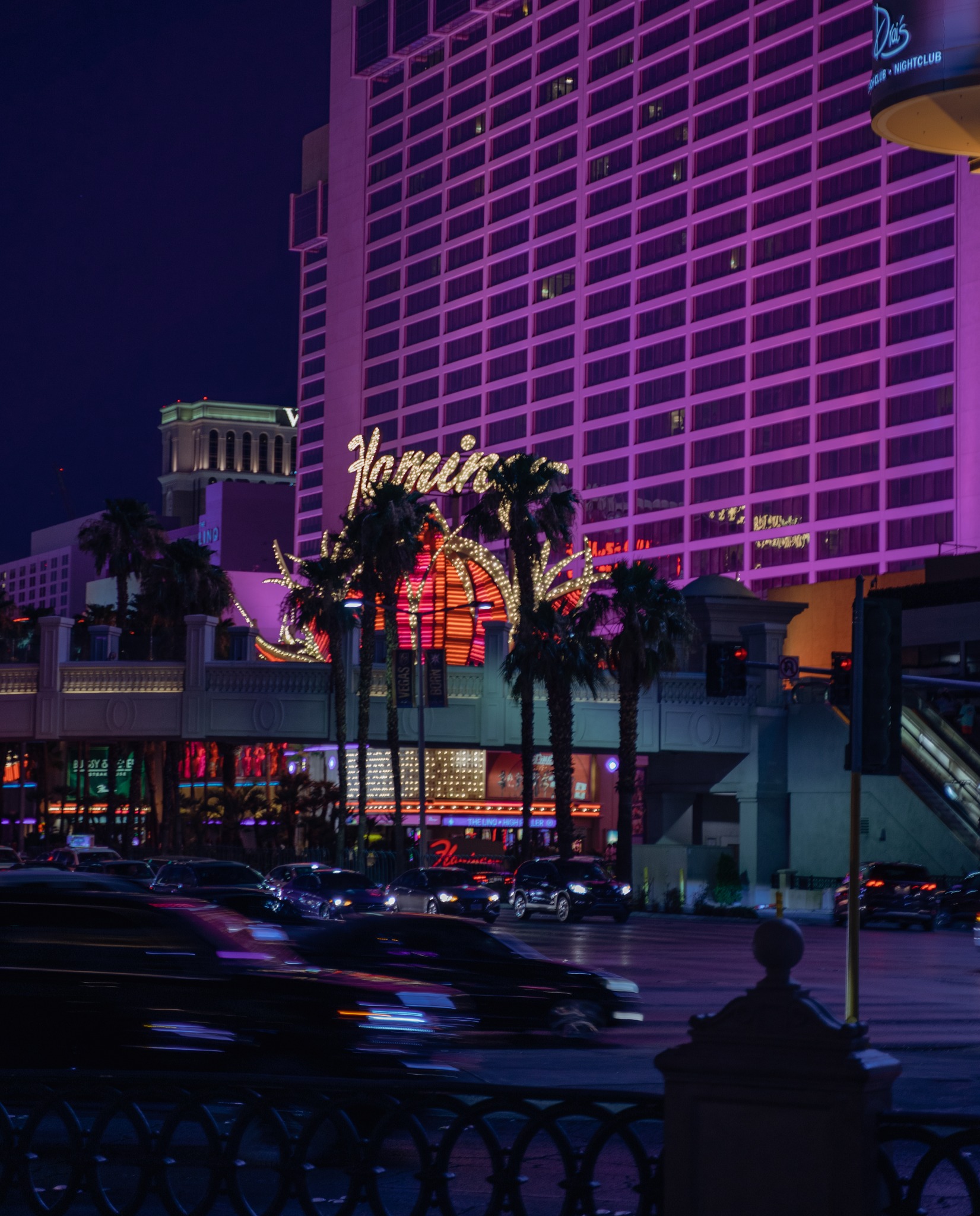 Nocny widok na hotel Flamingo w Las Vegas z neonowym napisem i palmami, rozmyte światła przejeżdżających samochodów.