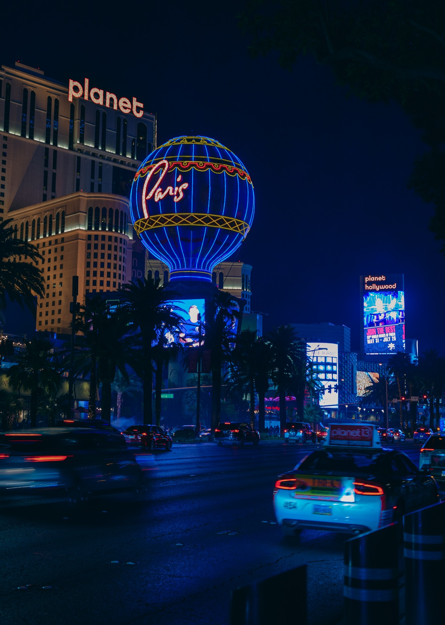 Nocny widok na Las Vegas Strip z neonami hoteli Planet Hollywood i Paris, z charakterystyczną wieżą Eiffla w kształcie balonu, ruch uliczny z rozmazanymi światłami samochodów, palmy i jasne reklamy.