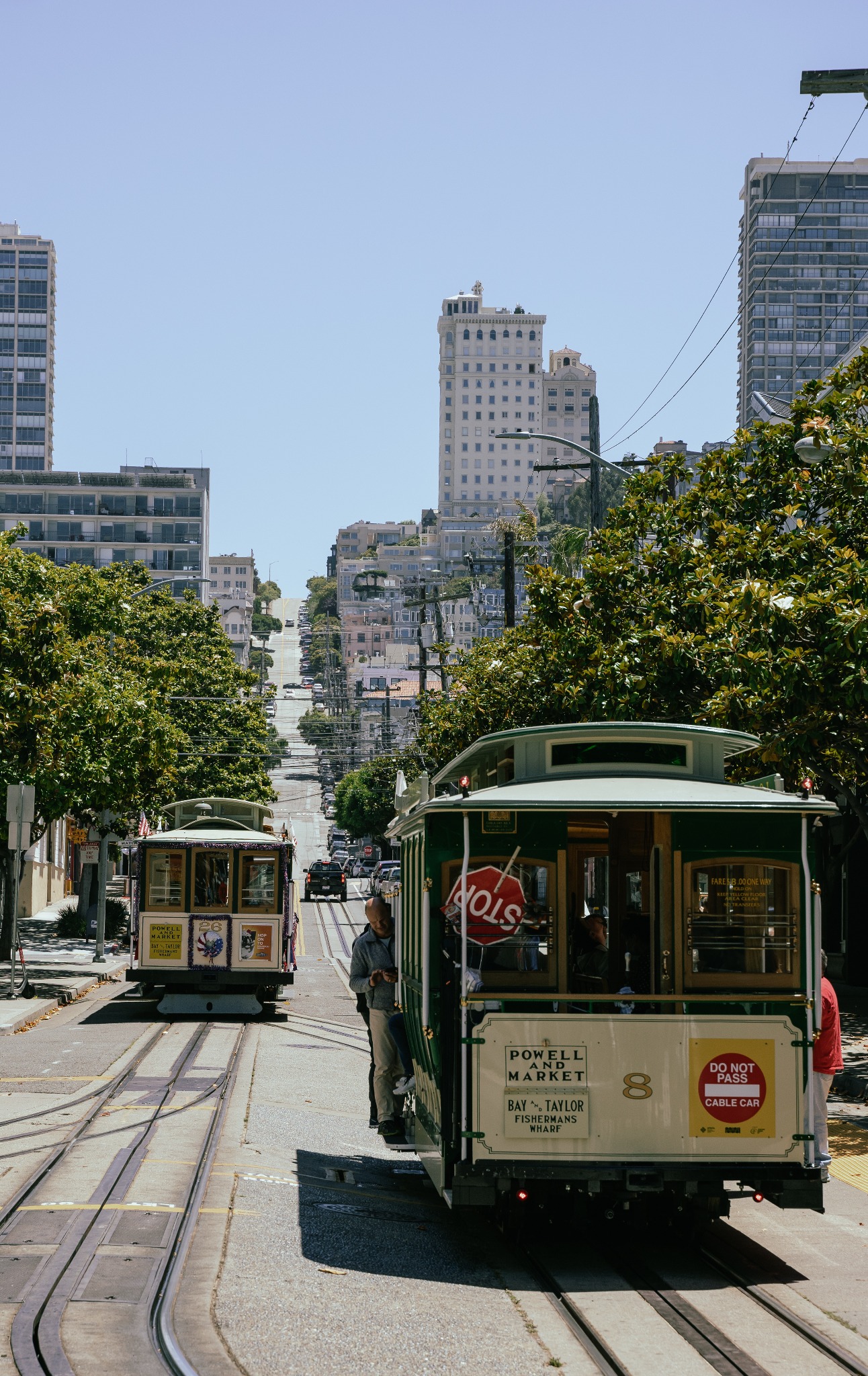 Dwa zielone tramwaje linowe w San Francisco, jeden z napisem 'Powell and Market' i numerem 8, jadące po torach w dół ulicy z wysokimi budynkami i stromym wzniesieniem w tle.