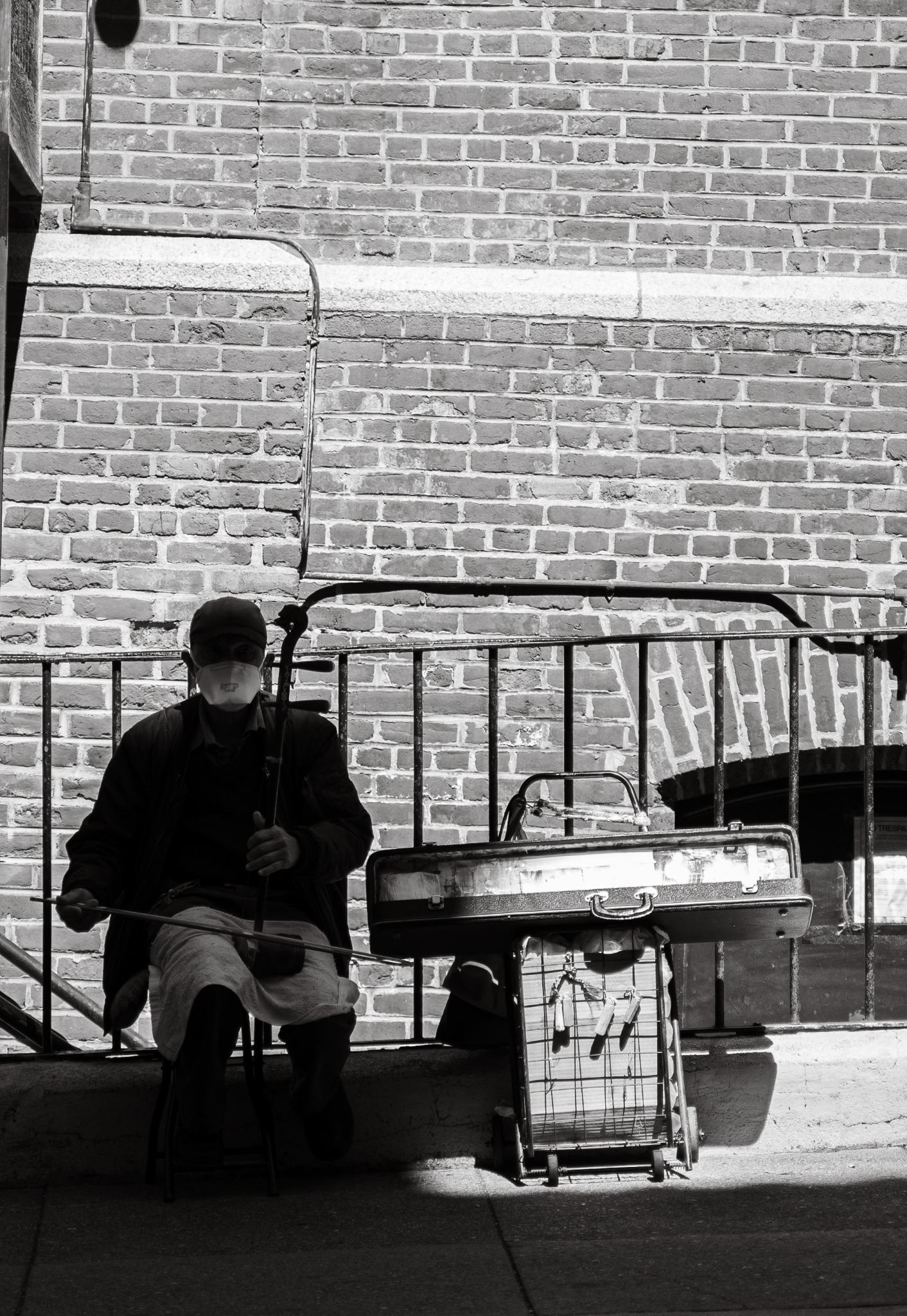 Czarno-białe zdjęcie ulicznego grajka z Poznania, siedzącego na krześle z chińskimi skrzypcami erhu, na tle ceglanej ściany i metalowej barierki.