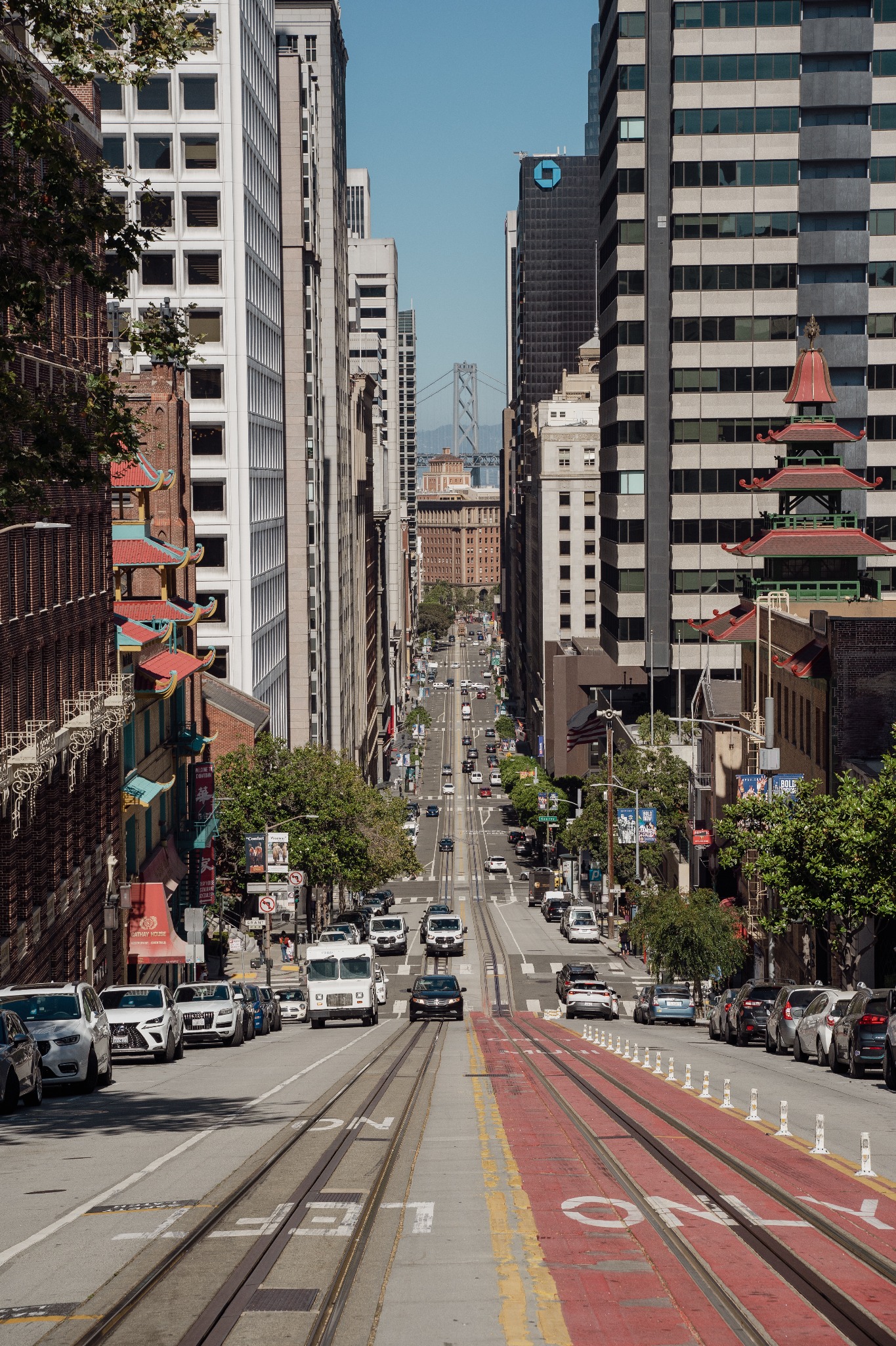 Widok z góry na ulicę w San Francisco z tramwajowymi torami, samochodami i drapaczami chmur w tle, w oddali widoczny most wiszący.