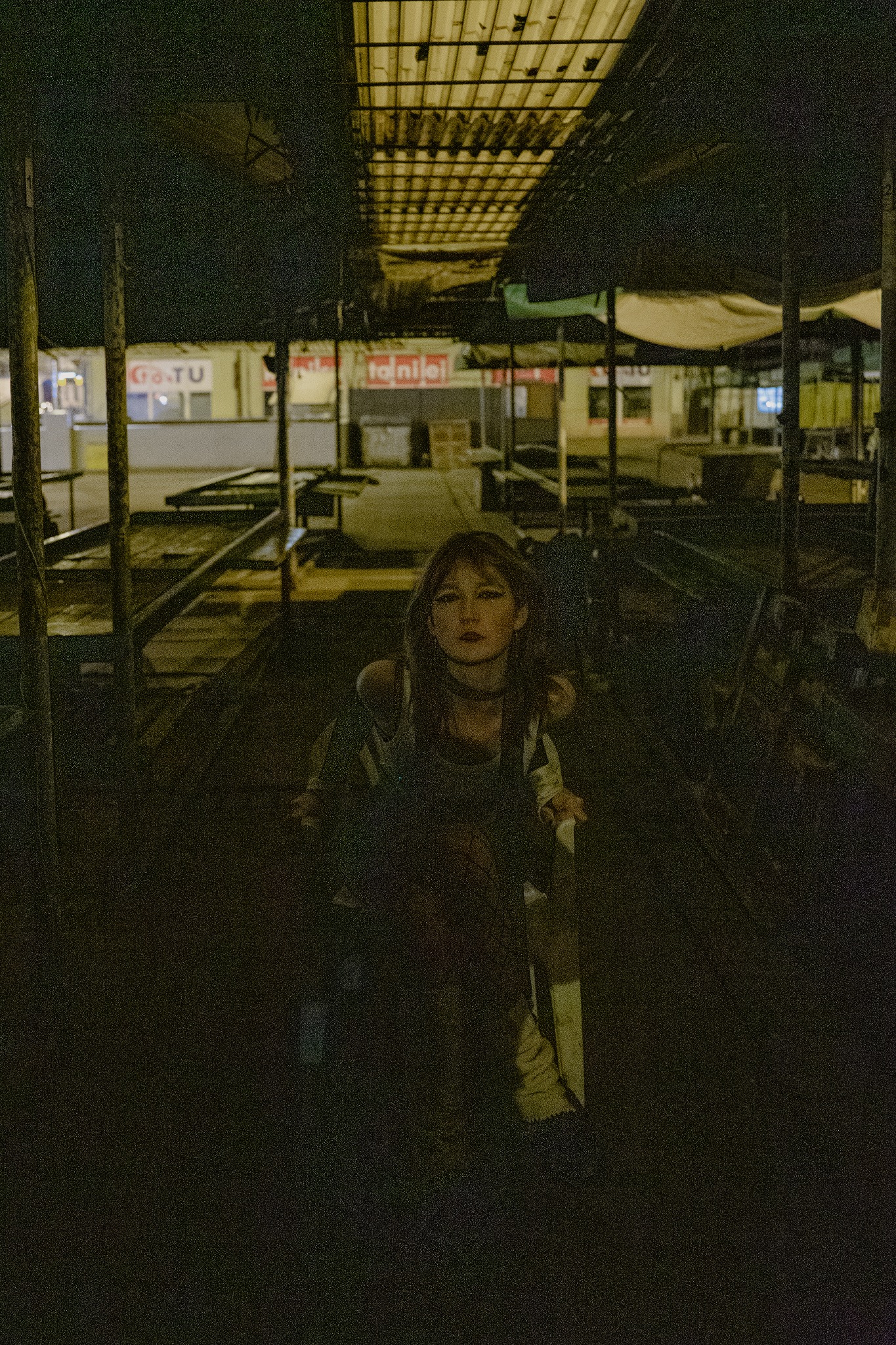 Portret kobiety z ciemnymi włosami i mocnym makijażem oczu w opuszczonej hali targowej z metalową konstrukcją dachu i pustymi stoiskami w tle, nocne oświetlenie.