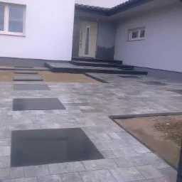 Ułożona kostka brukowa w odcieniach szarości z wstawkami z ciemnych płyt prowadząca do wejścia domu z białymi ścianami i szarymi elementami wykończeniowymi.