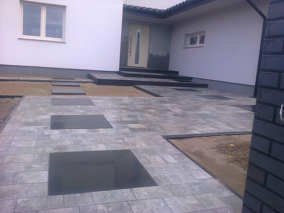 Ułożona kostka brukowa w odcieniach szarości z wstawkami z ciemnych płyt prowadząca do wejścia domu z białymi ścianami i szarymi elementami wykończeniowymi.