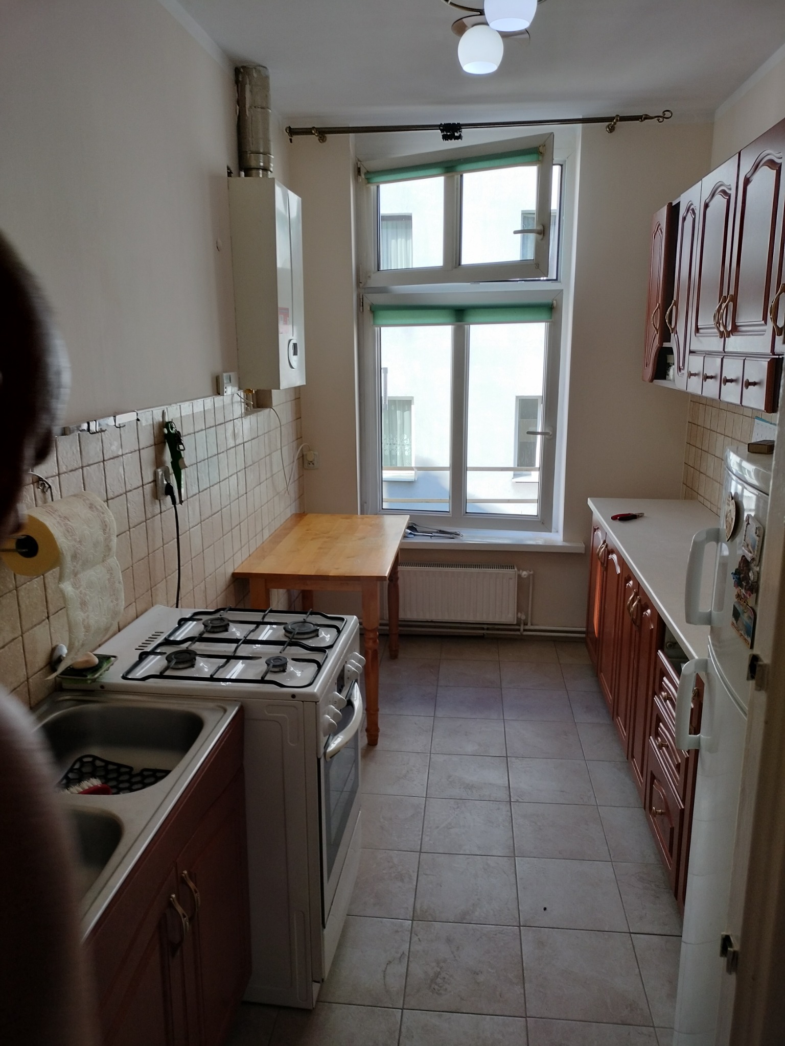 Wnętrze kuchni z beżowymi kafelkami, drewnianymi szafkami, białym piecem gazowym i stołem pod oknem z zielonymi roletami.