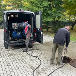 Pracownik w czerwonej kurtce i czapce pokazuje kciukiem do góry, drugi pracownik w czarnej bluzie czyści studzienkę kanalizacyjną za pomocą węża z wodą pod ciśnieniem, w tle widoczny samochód...