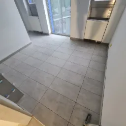 Świeżo ułożona, szara, kwadratowa podłoga z płytek w jasnym pokoju z dużymi oknami, widoczne narzędzie do rozprowadzania fugi i poziomice na ścianie.