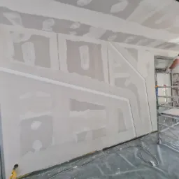 Prace przygotowawcze na ścianie z płyt kartonowo-gipsowych, widoczne łączenia i szpachlowanie, pracownik na rusztowaniu, zabezpieczona folią podłoga, dekoracyjne elementy na ścianie.