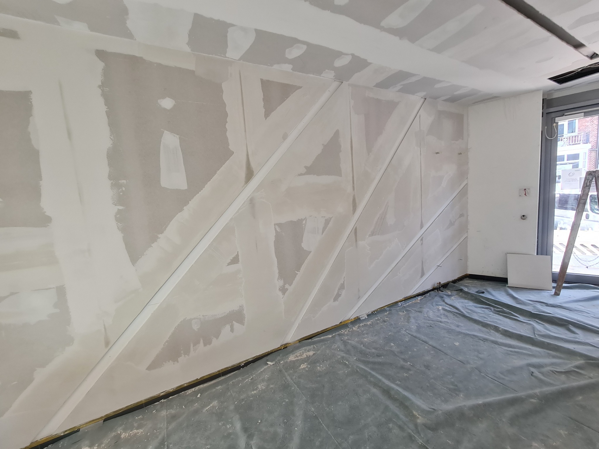 Ściana w trakcie szpachlowania z ukośnymi profilami, widoczne łączenia płyt kartonowo-gipsowych, podłoga zabezpieczona folią malarską, drabina oparta o ścianę przy oknie.