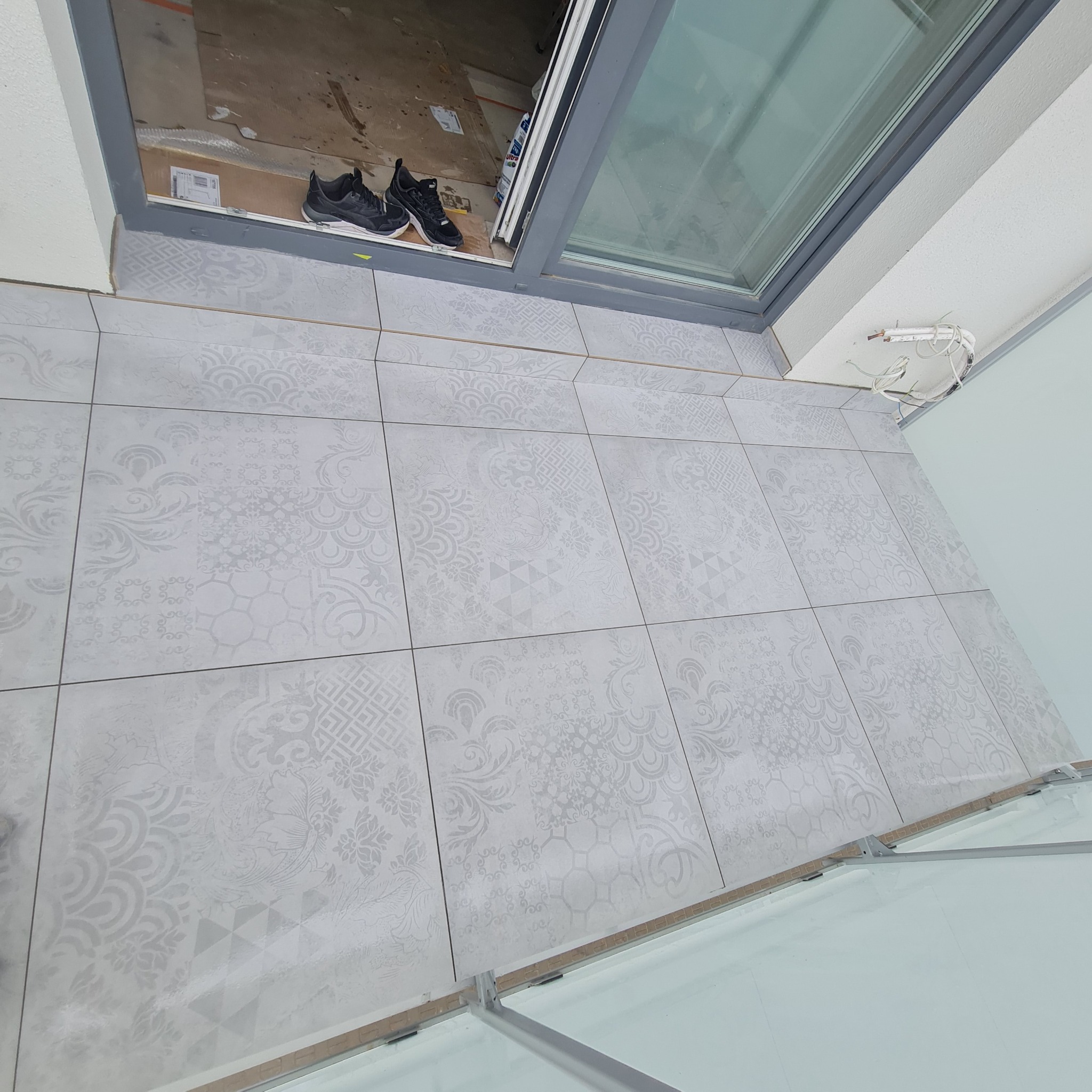 Ułożone szare płytki podłogowe z delikatnym wzorem na balkonie w Pułtusku, widoczne buty robocze i wystające kable.