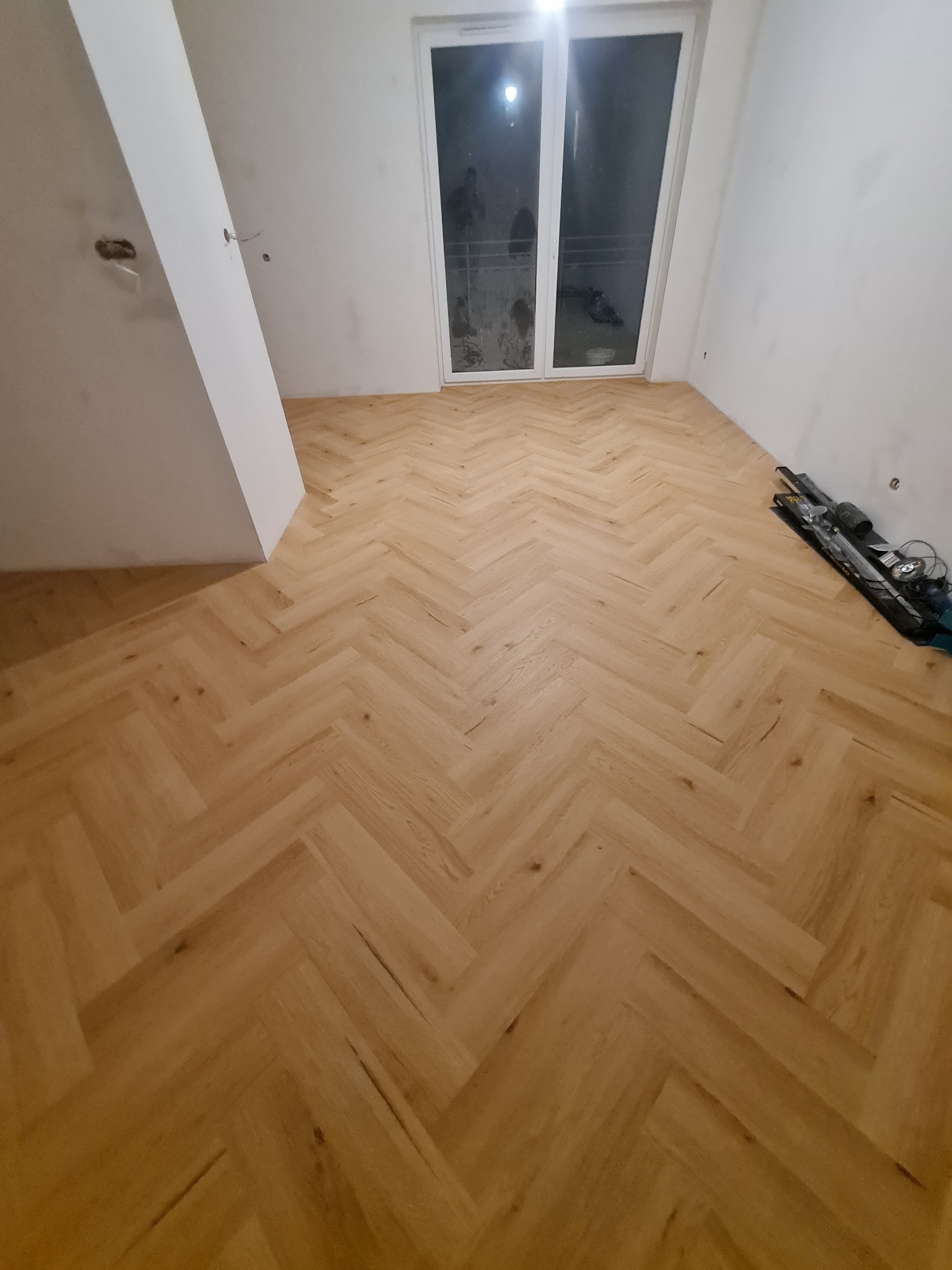 Parkiet ułożony w jodełkę w jasnym odcieniu drewna w pustym, odświeżonym pokoju z białymi ścianami, widoczne narzędzia glazurnicze przy ścianie.