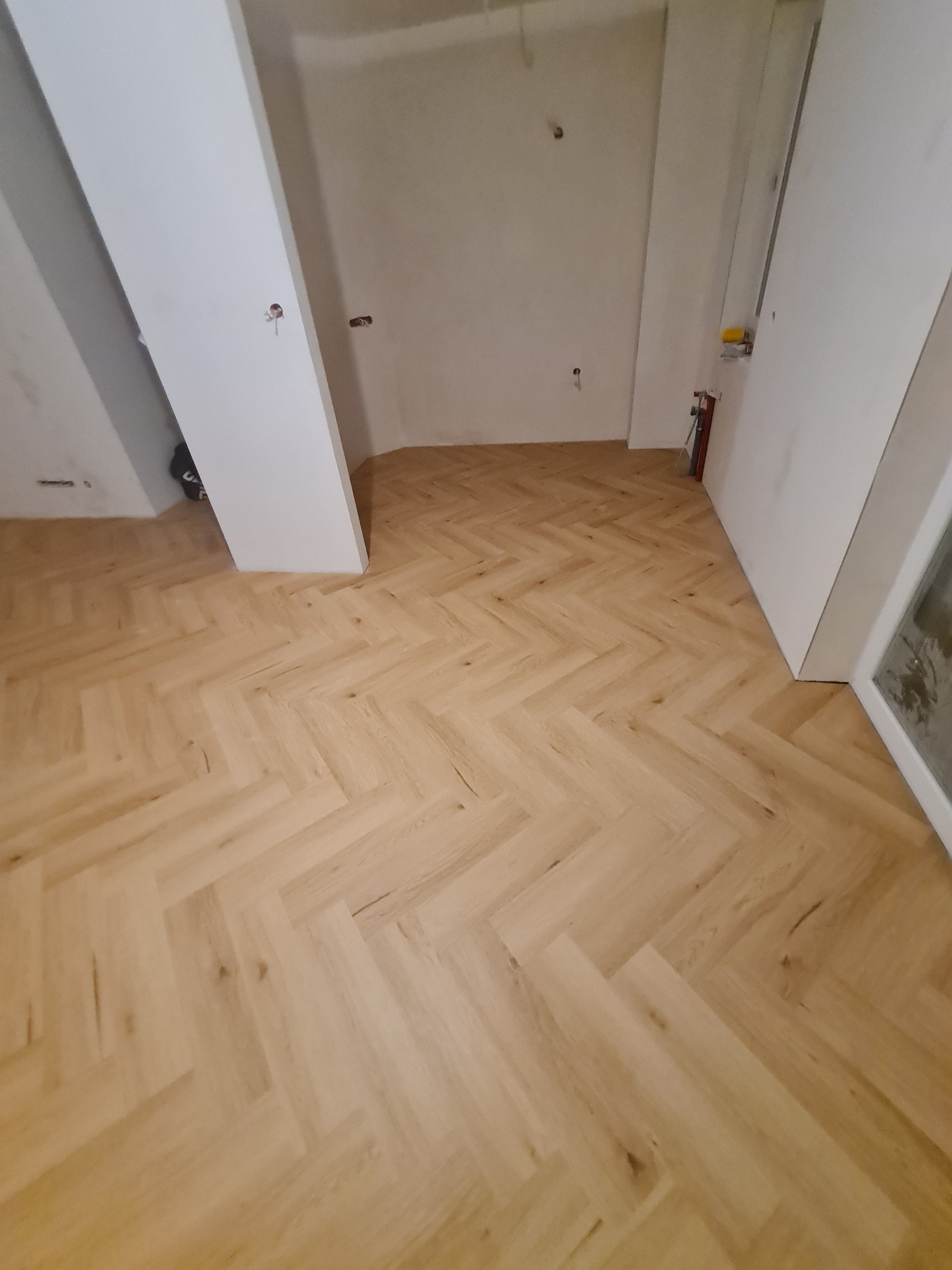 Parkiet ułożony w jodełkę w jasnym odcieniu, świeżo po położeniu, widoczne surowe ściany i przygotowane otwory na instalacje elektryczne, w tle fragment białych drzwi.