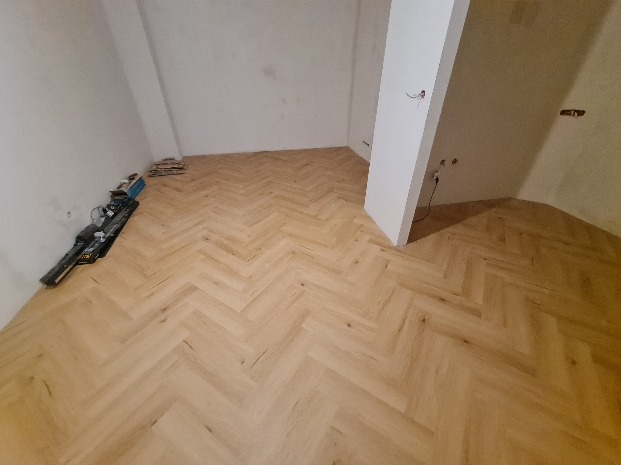 Parkiet ułożony w jodełkę francuską w jasnym odcieniu, widoczne narzędzia i materiały budowlane przy ścianie, surowy stan ścian i wystające kable elektryczne.