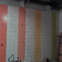 Ściana w trakcie układania płytek, z pionowymi pasami w kolorach białym, łososiowym, żółtym i oliwkowym, widoczne narzędzia glazurnicze na pierwszym planie, surowy stan wykończenia pomieszczenia.