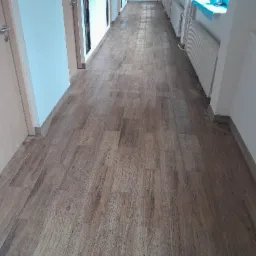 Długi korytarz z nowo ułożonymi panelami podłogowymi imitującymi drewno, widoczne drzwi po obu stronach i grzejniki pod oknami.