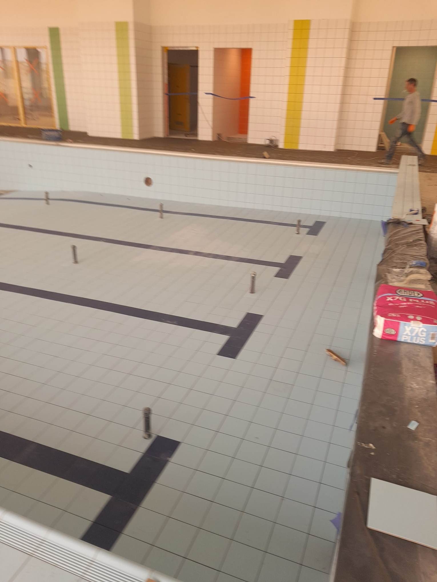 Wnętrze basenu w trakcie wykańczania: ułożone jasne płytki z ciemnymi pasami oznaczającymi tory pływackie, widoczny worek z klejem do płytek, fragment ściany z białymi płytkami i kolorowymi pasami...