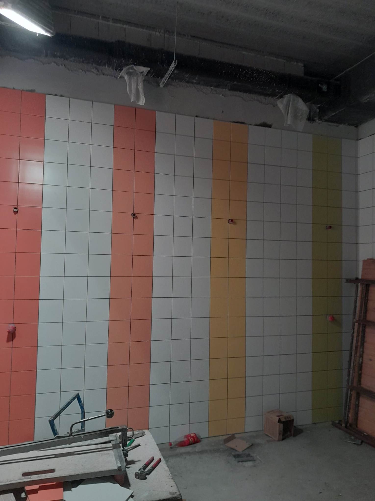 Ściana w trakcie układania płytek, z pionowymi pasami w kolorach białym, łososiowym, żółtym i oliwkowym, widoczne narzędzia glazurnicze na pierwszym planie, surowy stan wykończenia pomieszczenia.