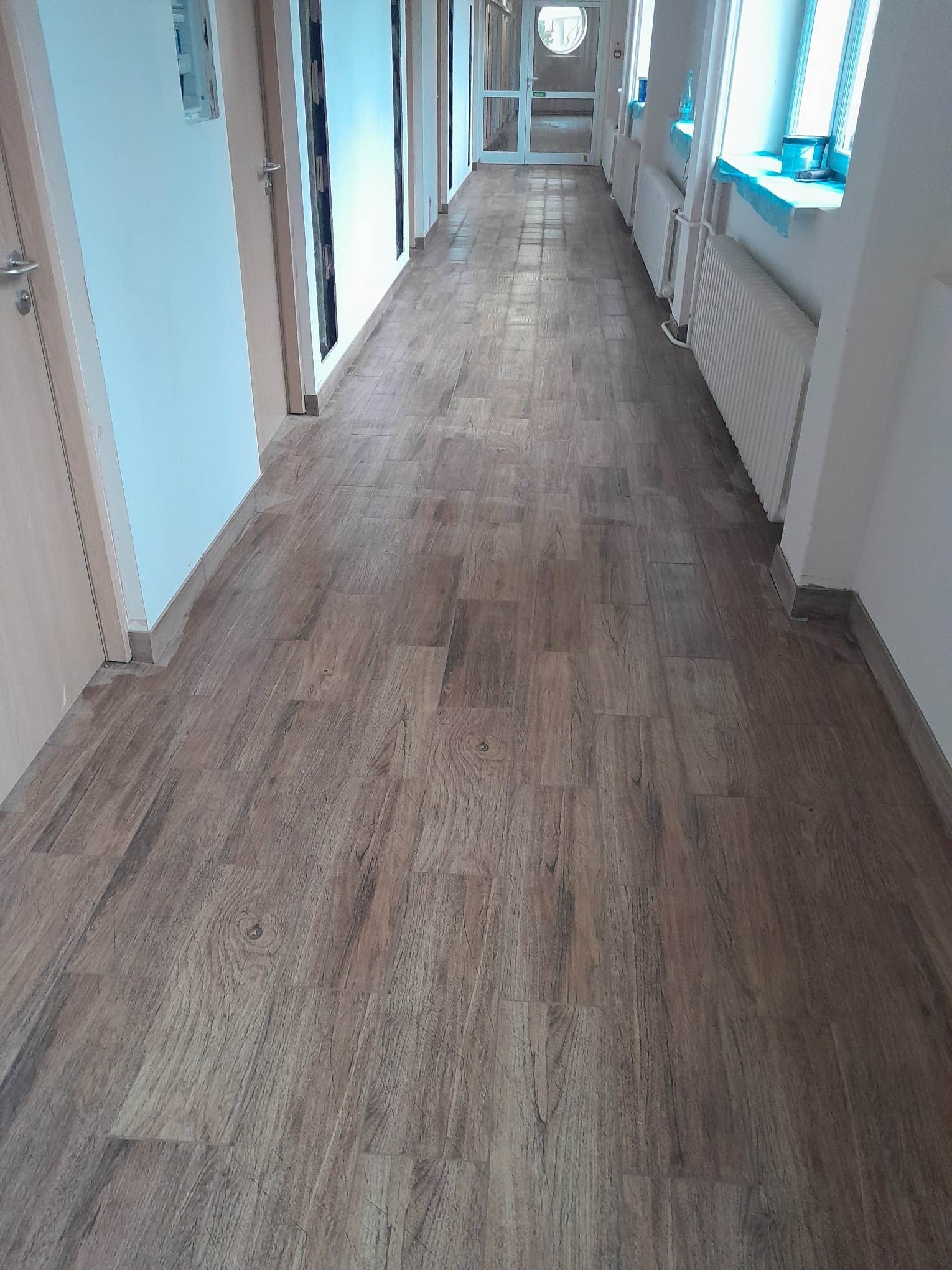Długi korytarz z nowo ułożonymi panelami podłogowymi imitującymi drewno, widoczne drzwi po obu stronach i grzejniki pod oknami.