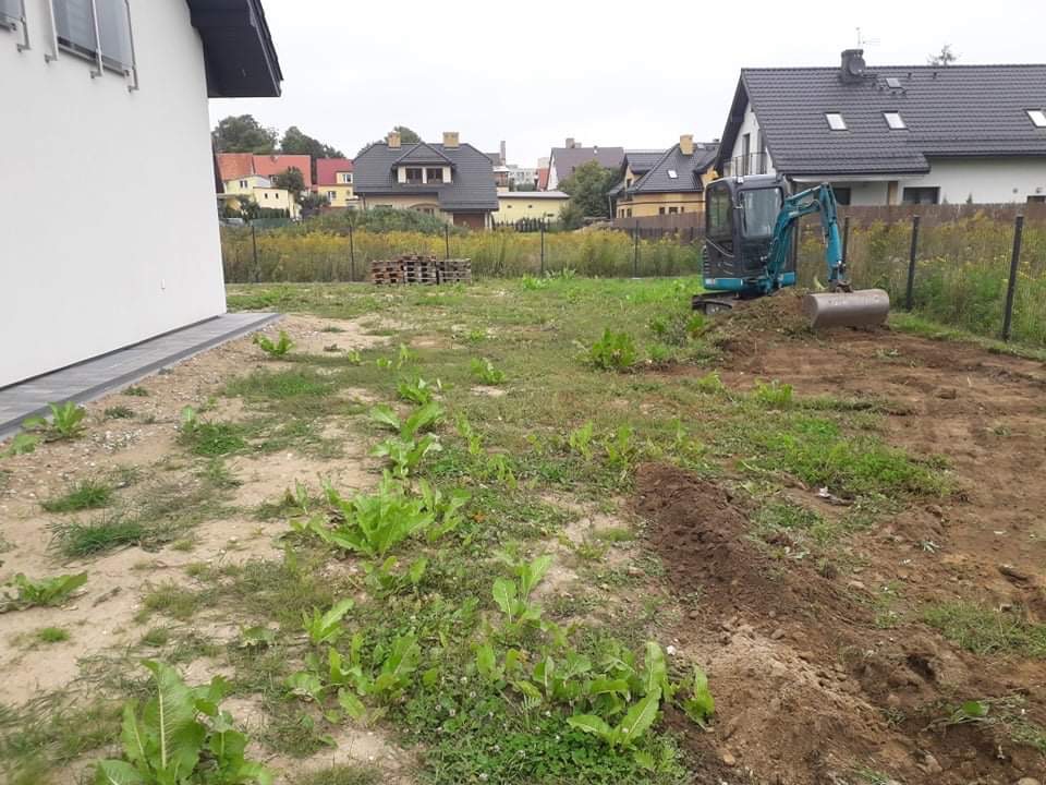 Mała, błękitna koparka wykonuje prace ziemne na działce przy nowym domu. W tle widoczne są inne budynki mieszkalne i palety z cegłami.