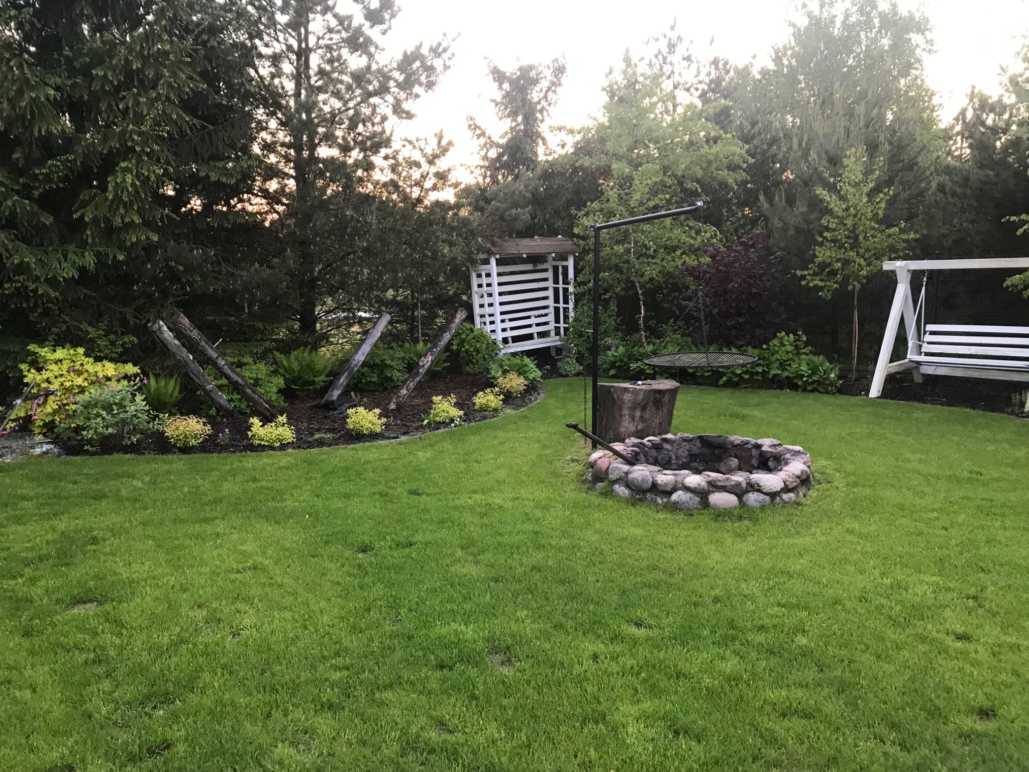 Utrzymany trawnik z kamiennym paleniskiem, drewnianą pergolą i huśtawką ogrodową w tle, otoczony drzewami i krzewami.