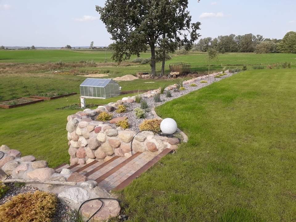 Ogród z kamienną kaskadą, schodami z kostki, rabatą z roślinami ozdobnymi i kulistą lampą, w tle szklarnia i zielone pola.