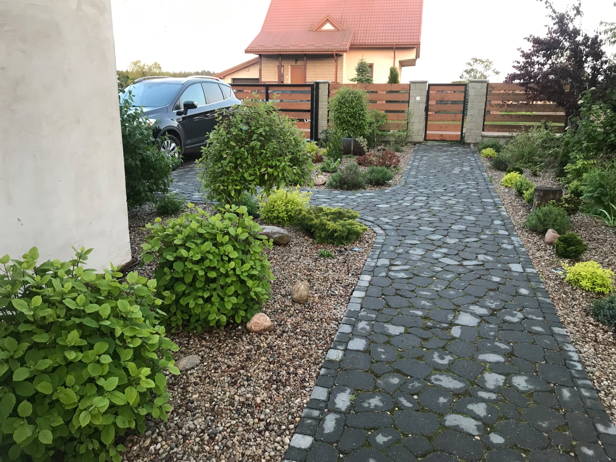 Ułożona z szarej kostki brukowej ścieżka prowadzi przez ogród z krzewami i drzewami, widoczny samochód osobowy i drewniany płot.