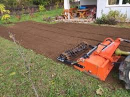 Mały, pomarańczowy traktor z wałem ugniata świeżo wyrównaną ziemię pod trawnik na działce przed domem. Widoczny fragment budynku z białym sidingiem i drewnianym tarasem.