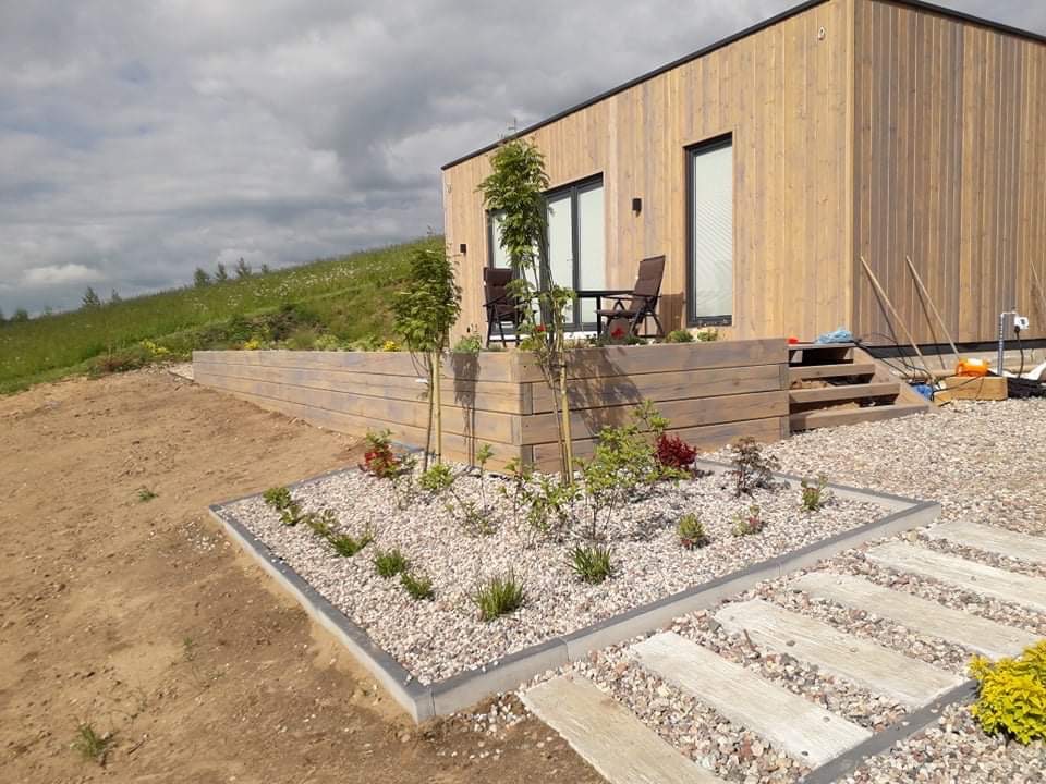 Nowoczesny dom z drewnianą elewacją i tarasem, ogródek z kamieniami i niską roślinnością, betonowy chodnik prowadzący do domu, widok na zielone wzgórze w tle.