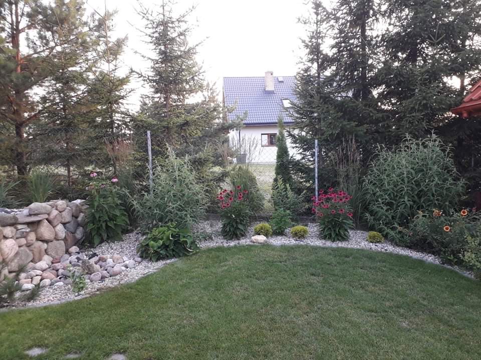 Ułożony z kamieni murek oporowy oddziela rabatę z iglakami, kwiatami i ozdobnymi trawami od trawnika. W tle widoczny dom z szarą dachówką i ogrodzenie z siatki.