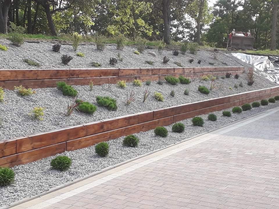 Ukształtowany teren w ogrodzie z drewnianymi belkami oporowymi, wysypany drobnym kamieniem i obsadzony roślinami. W tle widoczna maszyna budowlana.