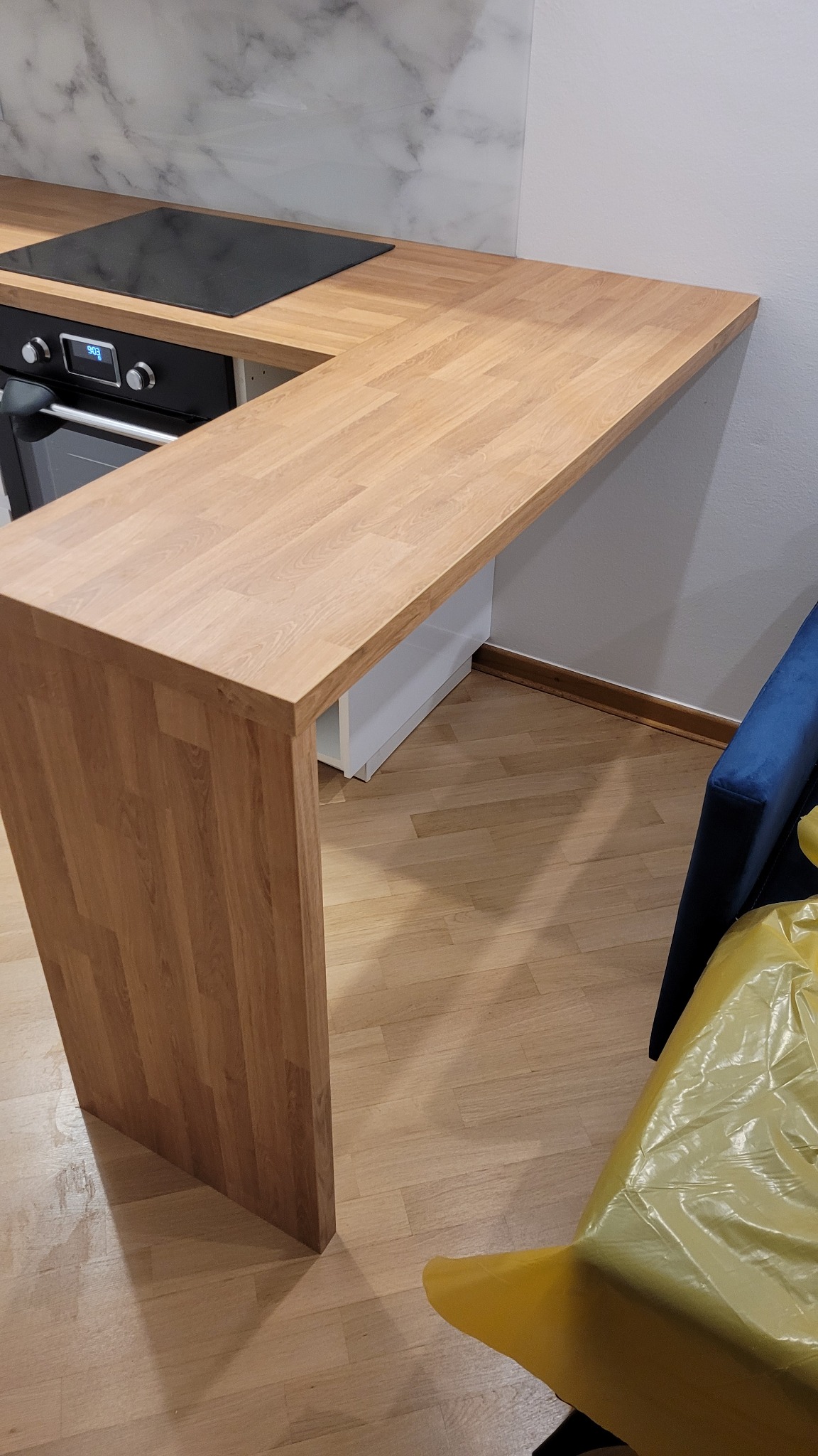 Fragment kuchni z blatem z drewna klejonego, wbudowaną płytą indukcyjną i piekarnikiem, widoczny fragment niebieskiego fotela z żółtą folią ochronną.