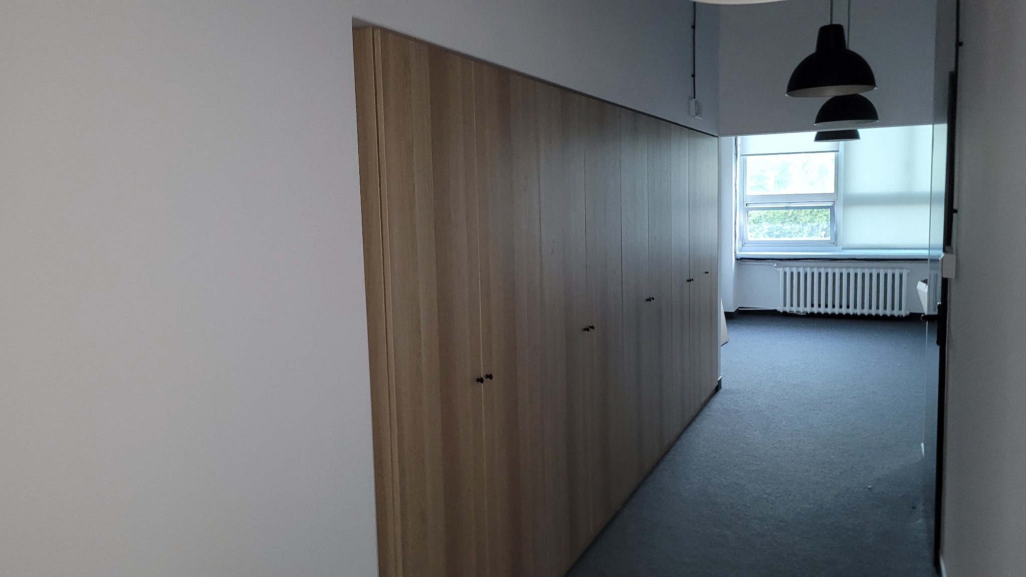 Długa, drewniana szafa wnękowa z ciemnymi uchwytami, ciągnąca się wzdłuż ściany korytarza, widok w głąb pomieszczenia z oknem i grzejnikiem.