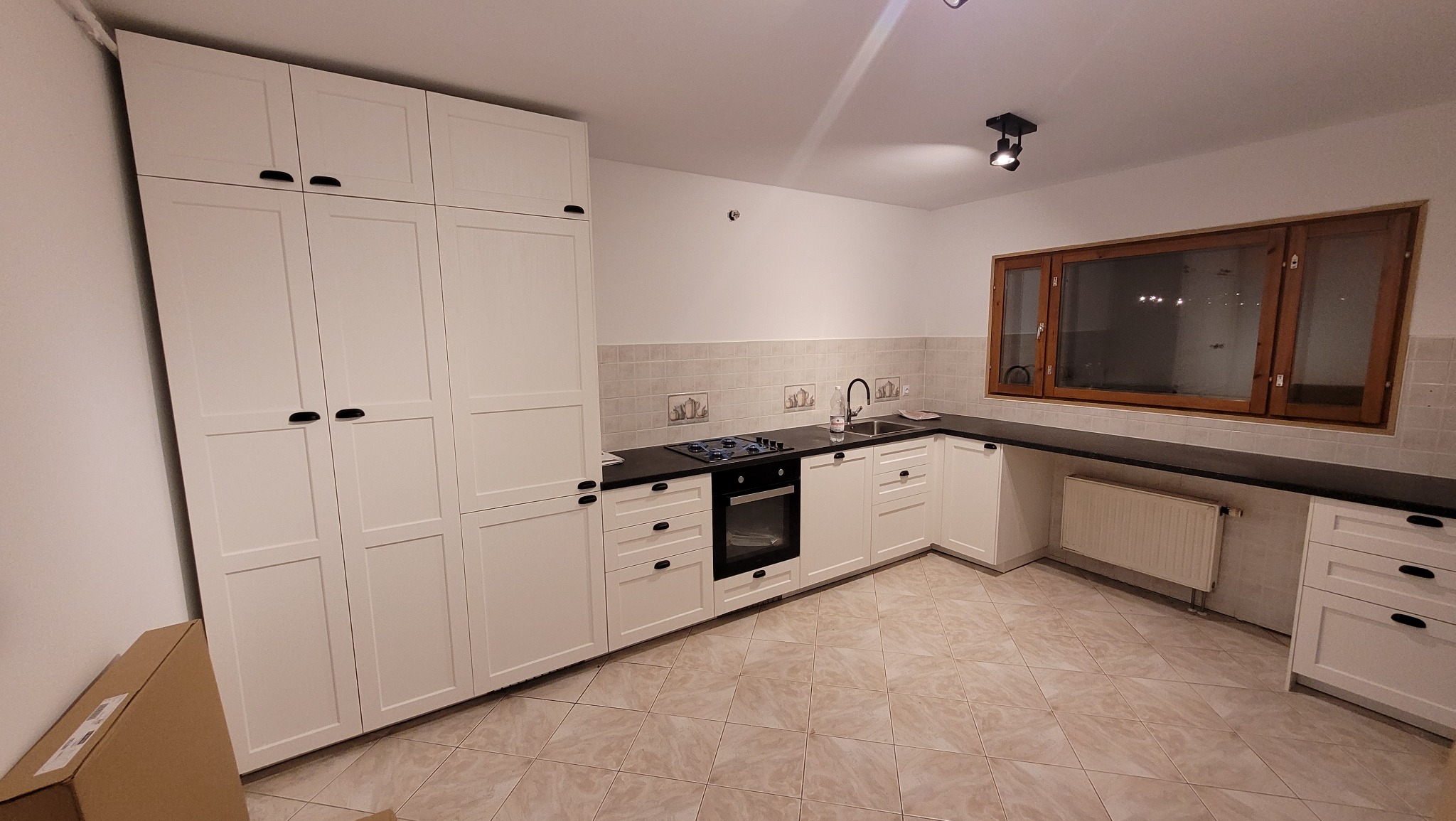 Jasna kuchnia z białymi szafkami i czarnym blatem, widok na całe pomieszczenie z perspektywy podłogi, płytki na ścianie, okno z drewnianą ramą, grzejnik pod blatem.