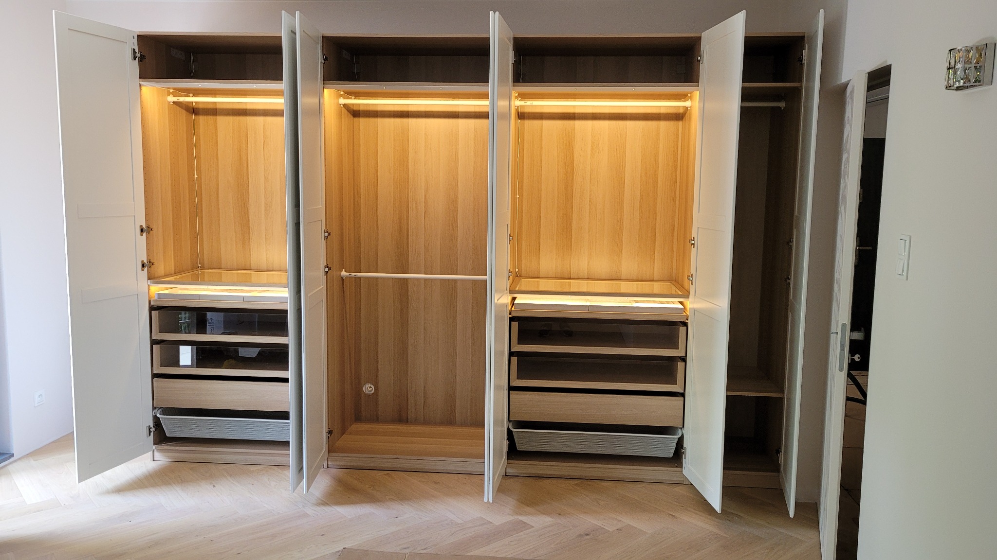 Wnętrze szafy z oświetleniem LED, drewnianym wykończeniem i szufladami z koszami, widoczne otwarte białe drzwi frontowe.