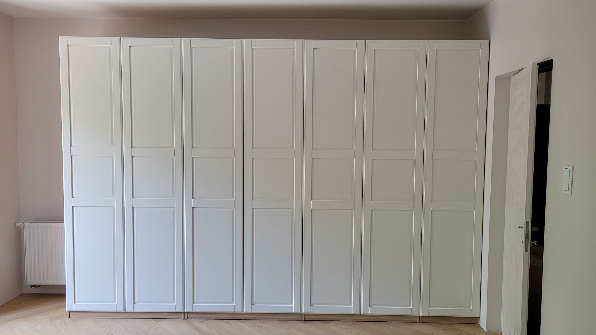 Biała, zabudowana szafa z ośmioma drzwiami panelowymi, osadzona na drewnianej podstawie, widoczna w jasnym wnętrzu z parkietem i grzejnikiem.