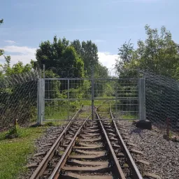 Metalowa brama z siatki z drutem kolczastym zamykająca tory kolejowe, widok z poziomu torów, zielone otoczenie.