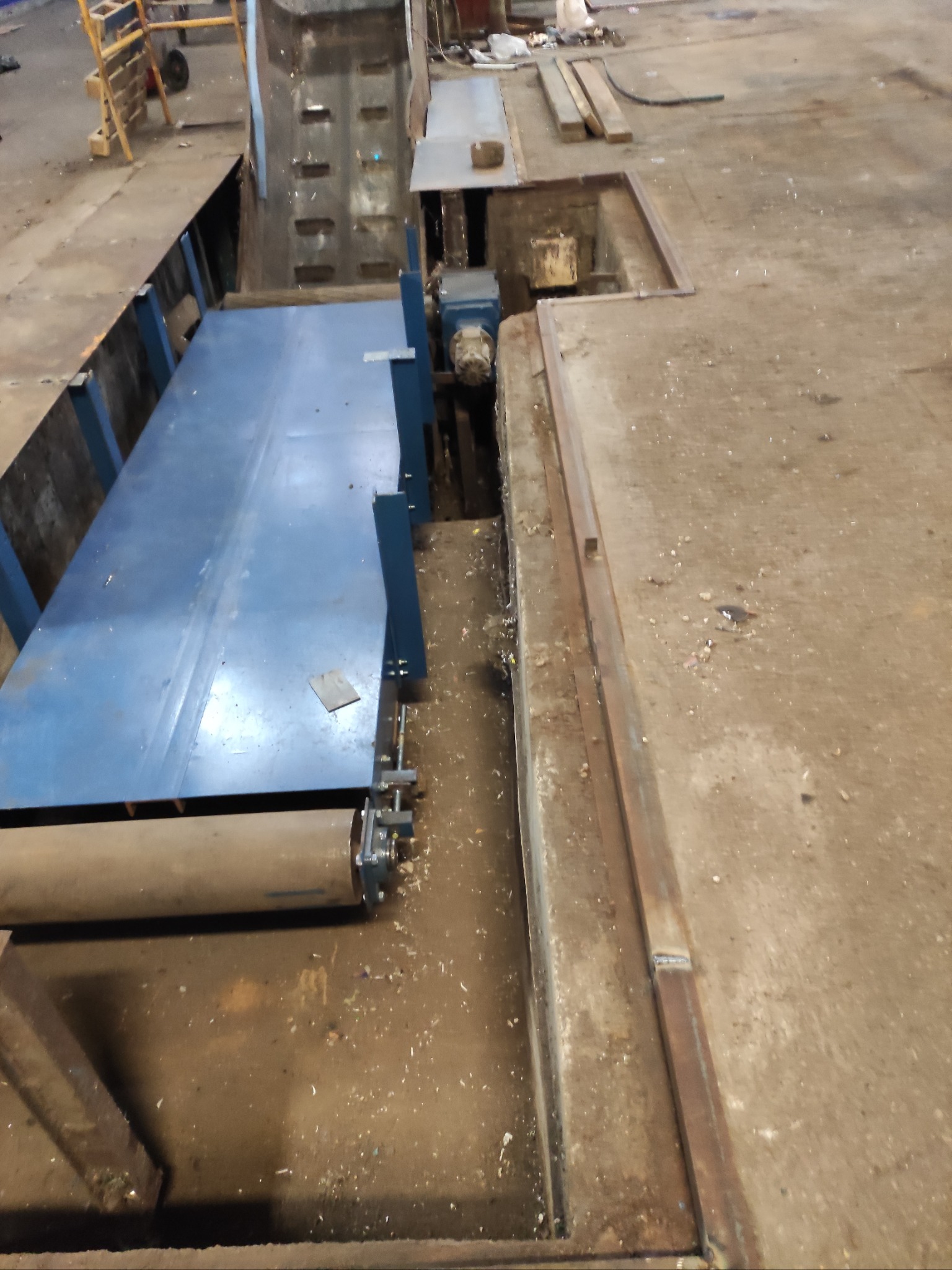 Nisko położony, niebieski przenośnik taśmowy z metalową konstrukcją w zagłębieniu podłogi hali przemysłowej, widoczne elementy mechaniczne i zabrudzenia.