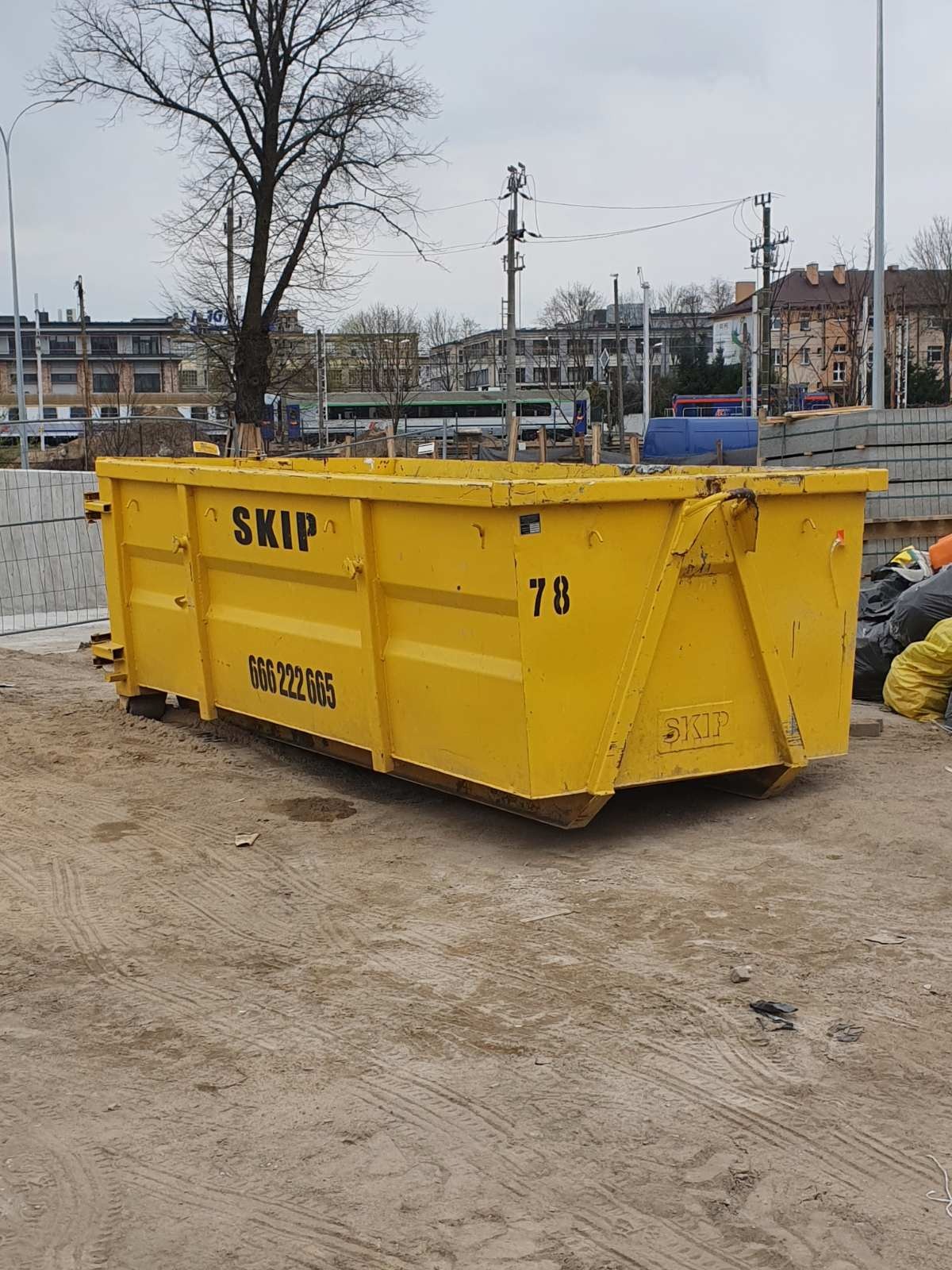 Żółty kontener SKIP z numerem 78 na placu budowy, widoczne ślady opon na piasku, w tle budynki i słupy energetyczne.