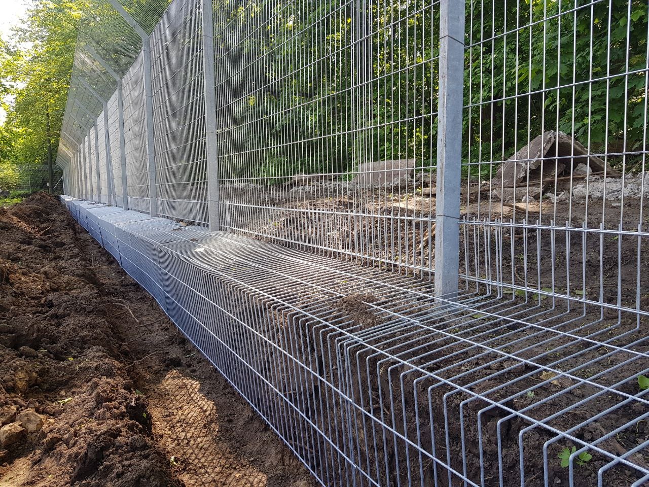 Długi, ocynkowany płot z siatki panelowej, częściowo wkopany w ziemię, z widocznymi słupkami i świeżo usypaną ziemią wzdłuż jego podstawy, na tle zieleni.