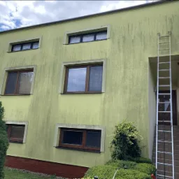 Dwupiętrowy budynek z żółtą elewacją pokrytą smugami zabrudzeń, z przystawioną aluminiową drabiną do wejścia.